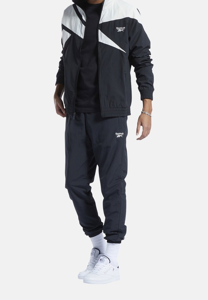 Man som bär en svart och vit Reebok träningsdräkt med logotyp, vita sockor av crew-modell och vita Reebok sneakers, står mot en enkel bakgrund.