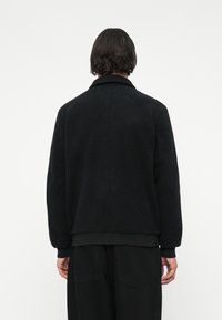 Giacca in pile nera con un esterno testurizzato, polsini e orlo a coste, e un colletto standard. Presenta un design semplice senza decorazioni.