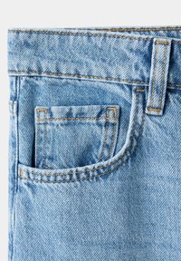 Gros plan sur un jean en denim bleu clair montrant une poche avant avec une couture orange détaillée et une passants de ceinture au-dessus.