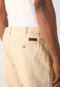 Gros plan d'une personne portant un pantalon en lin beige avec une poche arrière à bouton et une petite étiquette de marque en cuir au-dessus de la poche.