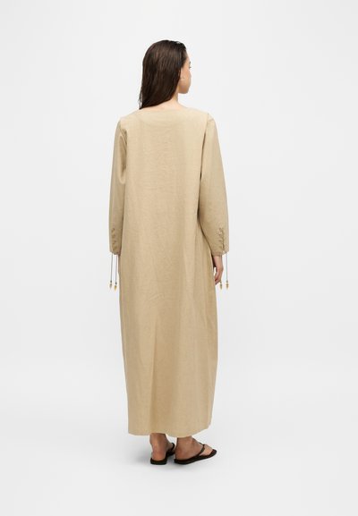 Femme aux longs cheveux portant une robe beige à manches longues et longue jusqu'aux chevilles, avec des sandales noires, dos tourné sur fond blanc.