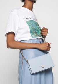 Jasnoniebieska torba na ramię z teksturowanego materiału, z klapką zapiętą na srebrny guzik. Regulowany pasek łańcuchowy zapewnia wszechstronność.