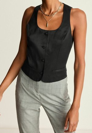 Femme portant un gilet noir sans manches boutonné, un pantalon gris à carreaux et des colliers dorés superposés.