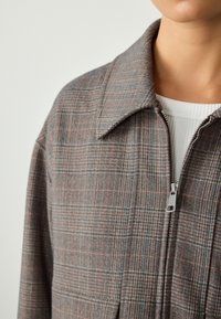 Giacca marrone a quadri con zip e colletto, caratterizzata da un tessuto leggermente strutturato e linee pastello a contrasto. Include dettaglio della tasca frontale.