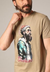 T-shirt en coton beige arborant un graphisme coloré d'un homme barbu portant des lunettes de soleil et un bonnet, avec le texte "Deeluxe."