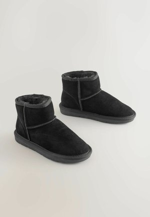 Černé semišové kotníkové boty s plyšovým podšívkou, kulatou špičkou a plochou gumovou podrážkou. Mají prošívané detaily a ohrnovací manžetu.