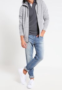 Šedá mikina na zip s kapucí přes tmavé polo tričko, modré džíny a bílé tenisky. Casual, uvolněný střih s ohrnutými manžetami.