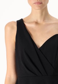 Musta hihaton mekko, jossa on syvä V-aukko, pehmeä kangas ja laskostettu muotoilu vyötäröllä. Sileä rakenne koko mekossa.