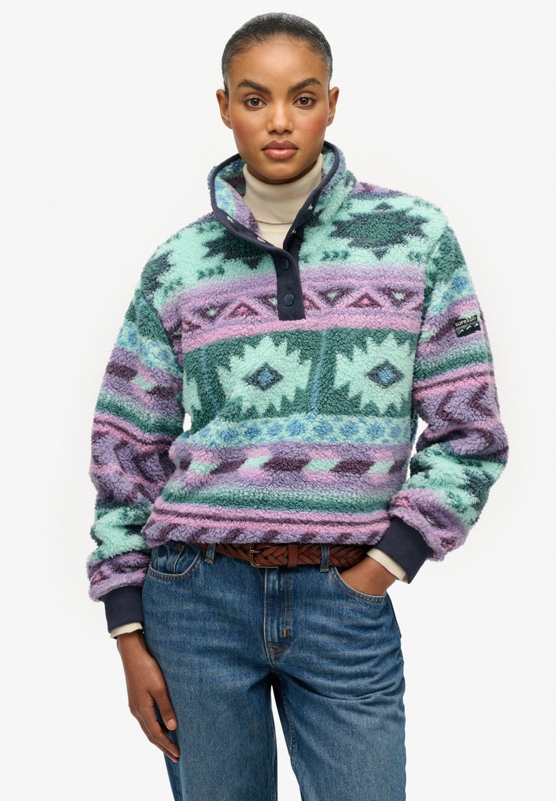 Fleece pullover in mintgroen, paars en blauw met geometrische patronen, voorzien van een hoge kraag, twee knopen en een marineblauwe manchetknoop. Draagt blauwe jeans.