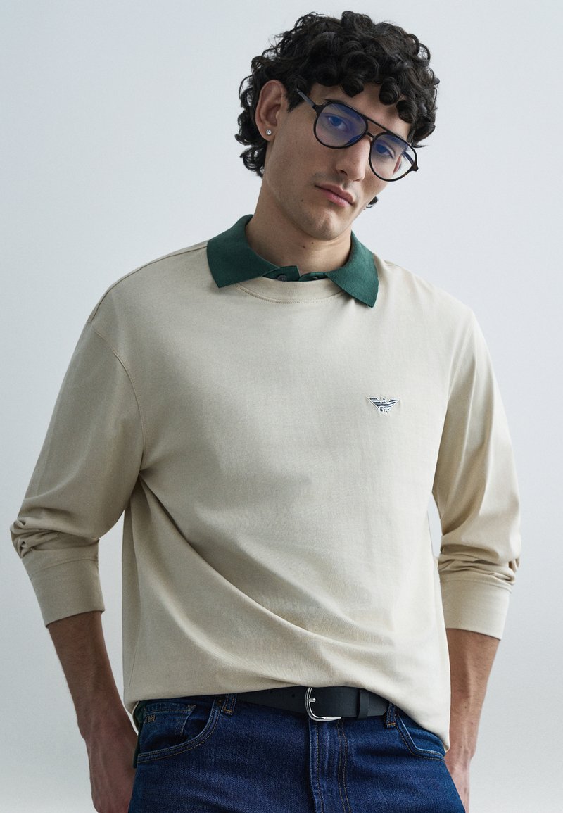 Bēša krekls ar garām piedurknēm un zaļu apkakli, ar mazu logo krūtīs, kombinēts ar tumši ziliem džinsiem.