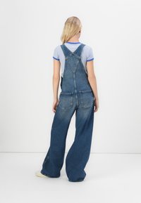 Dunkelblaue Denim-Latzhose mit lockerer Passform, gekreuzten Trägern, zwei Gesäßtaschen und weichem Material.