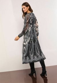 Rochie lungă alb-negru cu model paisley, mâneci lungi transparente și talie strânsă, asortată cu cizme negre până la genunchi.