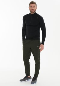 Zwart atletisch shirt met lange mouwen en halve rits, gecombineerd met olijfgroene tapered joggers met zijzakken en logo, gedragen met donkere sneakers.