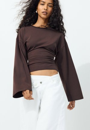Femme aux longs cheveux bouclés portant un haut moulant à manches longues marron foncé et un pantalon blanc taille haute sur un fond uni.