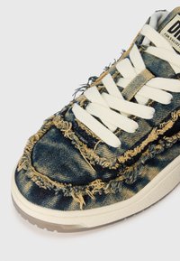 Dunker i denim med frynsete kanter, hvite snørebånd og en teksturert såle. Blå og gul fargekombinasjon med synlige sømdetaljer.