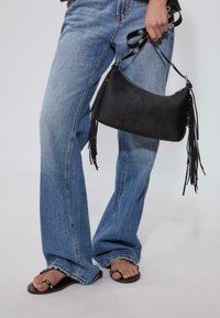 Sort faux suede taske med frynser, buet form og lynlåslukning, holdt af en person iført lyseblå brede bukser og sorte flip-flops.