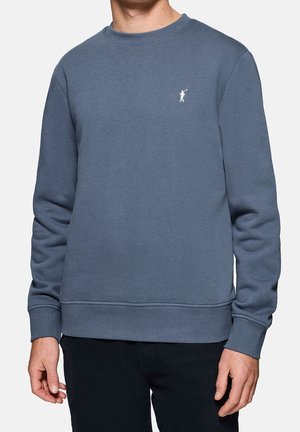 Mies pukeutuneena yksinkertaiseen siniseen pyöreän kaula-aukon collegepaitaan, jossa on pieni valkoinen Polo-pelaajan logo rinnassa, yhdistetty mustiin housuihin.