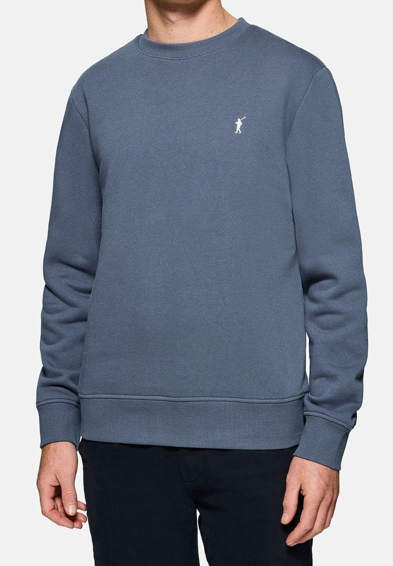 Homme portant un sweat-shirt bleu uni à col rond avec un petit logo blanc de joueur de polo sur la poitrine, associé à un pantalon noir.