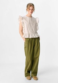 Lys beige bluse med flæser på ærmerne og knaplukning, sat sammen med løse olivengrønne bukser, hvilket skaber en afslappet silhuet.