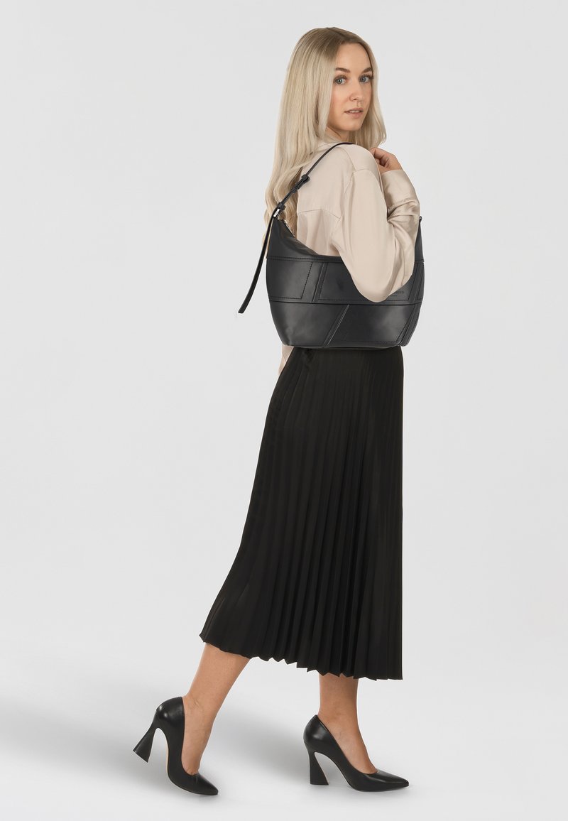 Sac hobo en cuir noir avec une forme arrondie, une anse plate et des détails de couture visibles. Associé à une jupe plissée noire et des escarpins.