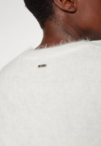 Gros plan sur le cou et le haut du dos d’une personne portant un pull blanc doux avec un petit logo métallique « BOSS » près du col.