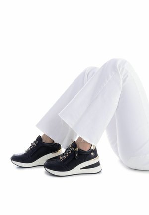 Chaussures de sport noires en mesh et matériaux synthétiques, accents métalliques, semelle blanche, et design à lacets, associées à un pantalon blanc évasé.