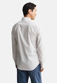 Mand med ryggen mod, iført en hvid langærmet skjorte og blå jeans mod en ensartet lys baggrund.