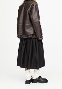 Veste en cuir marron avec un col en fourrure, superposée à une chemise à carreaux, associée à une jupe noire, des chaussettes blanches montant jusqu'aux genoux et des chaussures noires.