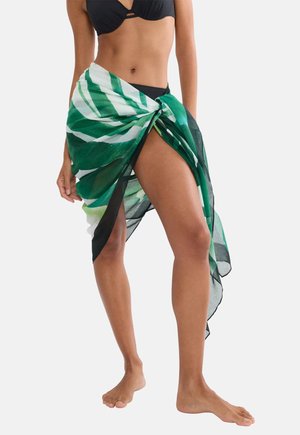 Kobieta w czarnym bikini z zielono-białym, wzorzystym sarongiem zawiązanym w talii, stojąca boso na jednolitym tle.