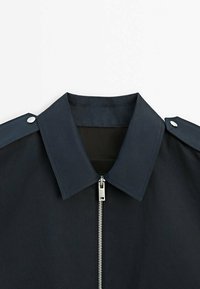 Granatowa kurtka z wyraźnym kołnierzem, srebrnym zamkiem błyskawicznym i guzikami na ramionach, wykonana z gładkiego, teksturowanego materiału.