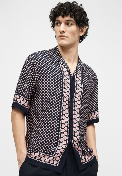 Άνδρας με σγουρά μαλλιά, φορώντας μπλε πουκάμισο με σχέδιο σειρών από ροζ καρδιές, στέκεται με τα χέρια στις τσέπες, κοιτάζοντας προς τα πλάγια.