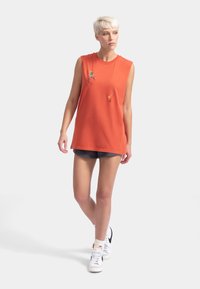 Débardeur sans manches en coton orange avec des motifs brodés colorés, associé à un short noir et des baskets montantes blanches.