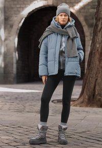Veste matelassée bleu clair avec capuche, associée à une écharpe en maille grise et un bonnet. Leggings noirs et bottes grises isolées à lacets complètent la tenue.