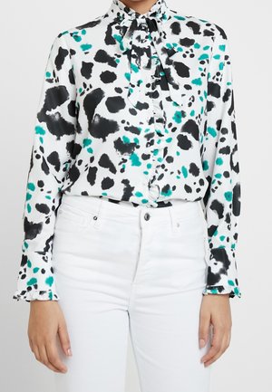 Femme portant un chemisier blanc avec des taches abstraites noires et turquoise, des poignets à volants et un col à nœud, associé à un pantalon blanc taille haute.