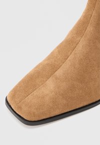 Gros plan sur une botte pointue en daim beige avec des coutures visibles et une semelle noire sur fond blanc.