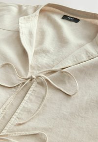 Lichtbeige shirt met een kraag, gekenmerkt door een gestrikte taille en een zachte, gladde textuur. Merkenlabel is zichtbaar in de halslijn.