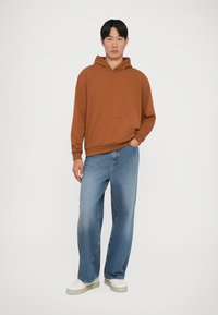 Hanorac maro cu buzunar frontal, asortat cu blugi largi de culoare albastru deschis și adidași albi. Material neted, cu o croială lejeră.