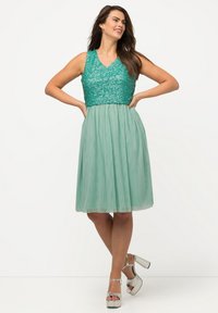 Vestido de lentejuelas en color verde azulado con un corsé ajustado y una falda fluida, combinado con tacones de plataforma plateados. El vestido presenta un escote en V y un diseño sin mangas.