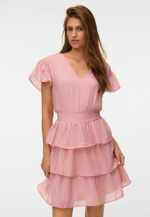 Mujer con cabello largo y castaño que lleva un vestido rosa claro con volantes y mangas acampanadas, de pie sobre un fondo blanco liso.