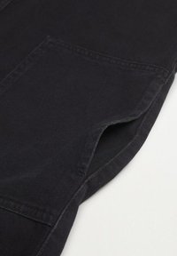 Gros plan sur le bas de jambe d'un pantalon en tissu noir avec des coutures visibles et un ourlet légèrement replié sur fond blanc.