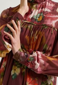 Een hand rust op de mouw van een bordeauxrood bloemenjurk, met levendige bloemen en een ritsdetail op de borst.