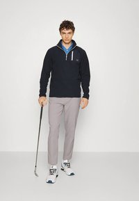 Giovane uomo in piedi con abbigliamento da golf casual, che tiene un putter da golf, indossando un pullover nero, pantaloni grigi e scarpe da ginnastica bianche su sfondo semplice.