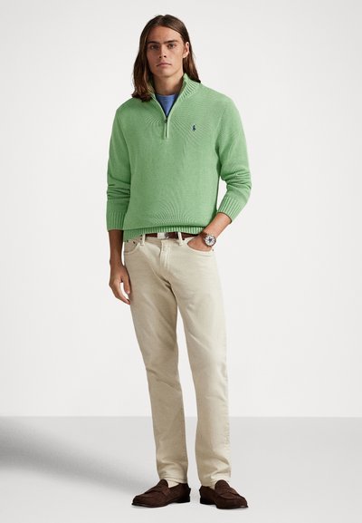 Zöld kötött pulóver félcipzáros gallérral, kék ing felett viselve. Világos bézs nadrággal és sötétbarna cipővel párosítva. Minimalista stílus.