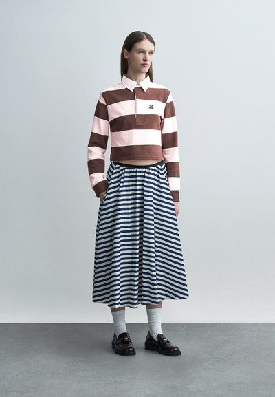 Γυναίκα που φοράει κοντό ροζ και καφέ ριγέ μπλουζάκι με μακριά μανίκια, μπλε και λευκή φούστα μέσης με διαγώνιες ρίγες, λευκές κάλτσες και μαύρες μοκασίνες.