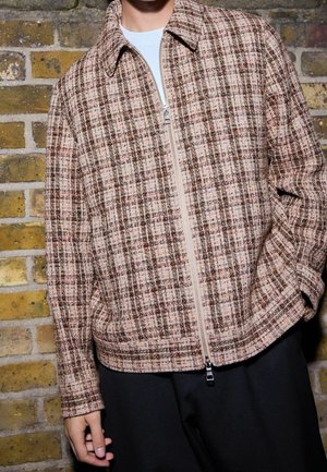 Osoba ubrana w brązowo-beżową, kratowaną kurtkę na zamek błyskawiczny, na białej koszulce, stojąca na tle żółto-szarej ściany z cegieł.