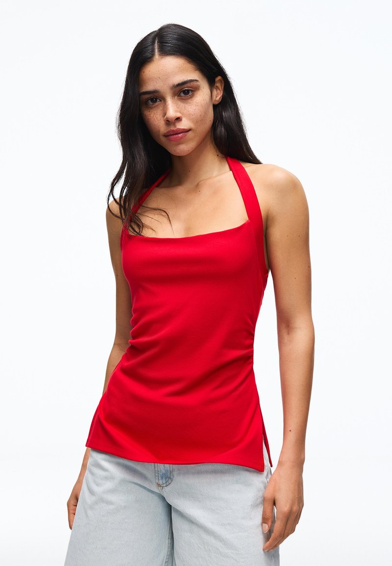 Femme aux longs cheveux foncés portant un haut dos-nu rouge et un jean bleu clair, debout devant un fond blanc uni.