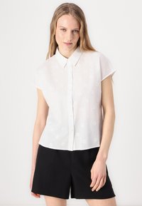 Camicetta bianca a maniche corte con colletto, caratterizzata da motivi floreali ricamati. Il materiale sembra leggero e semi-trasparente.