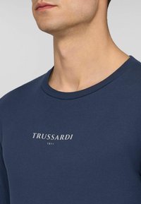 Zbliżenie na osobę w granatowej koszuli z napisem "TRUSSARDI 1911" w białym kolorze na piersi.