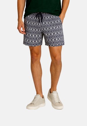 Mężczyzna w granatowo-białych szortach ze sznurkiem, czarnej koszulce i beżowych sneakersach, stojący z jedną ręką w kieszeni.