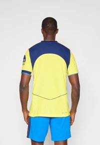 Sports trøje med en skarp gul krop og marineblå detaljer samt et tekstureret design, parret med blå shorts.